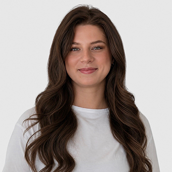 Woman with long wavy brown hair, wearing a white shirt, smiling against a light background.